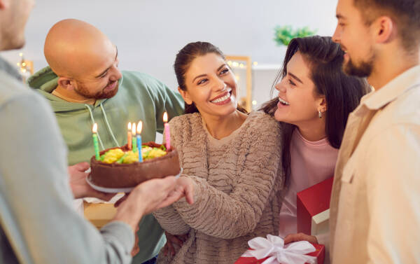 A family celebrates a birthday indoors, passing a birthday cake to a young woman as everyone smiles.