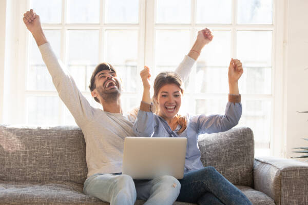 A happy young couple celebrates an online victory, raising their hands and screaming with joy as they look at the laptop screen together.