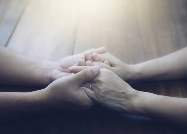 A husband and wife hold hands and pray together at a wooden table, illuminated by light from above.