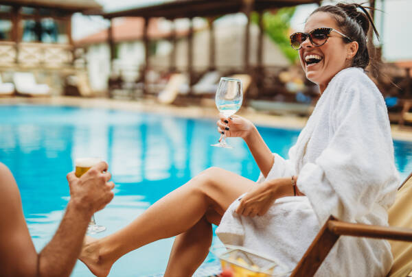 A laughing 29-year-old woman sits by the pool on vacation with her husband, wearing a robe.