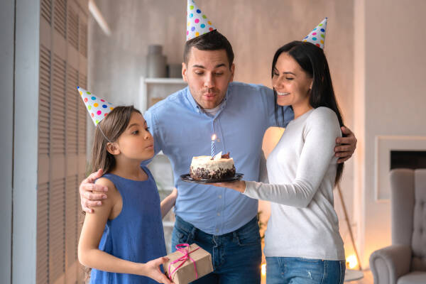 A man blows out a candle on a cake at home next to his wife and young daughter who holds a gift, all wearing party hats.