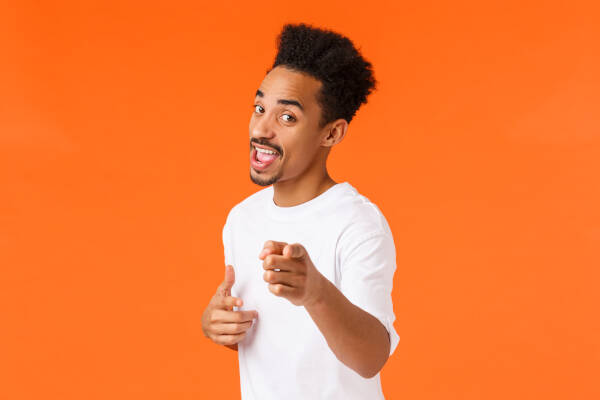 A man in a white t-shirt pointing at the camera as he stands against an orange background.