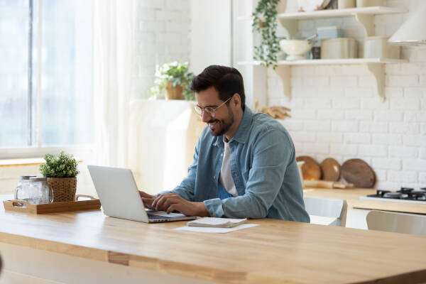 A man in glasses and a denim shirt sits at a kitchen island, smiling while typing on a laptop in a bright, plant-filled kitchen.