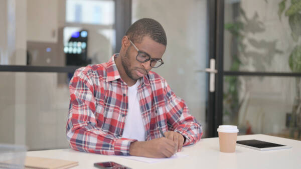 A man in glasses and a red plaid shirt writes on paper at a table in an office, with a coffee cup and tablet beside him.