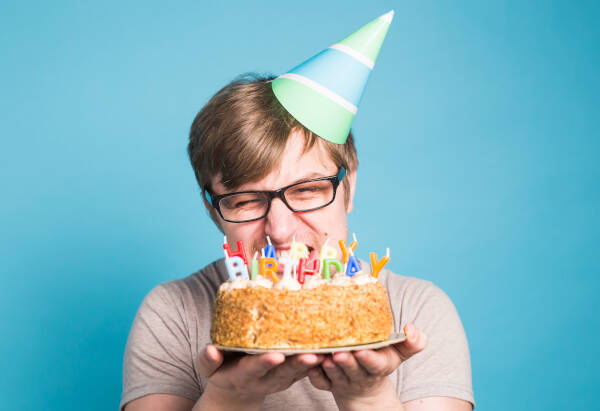 A man in spectacles and a party hat holds up a cake and pulls a strange expression, against a light blue background.