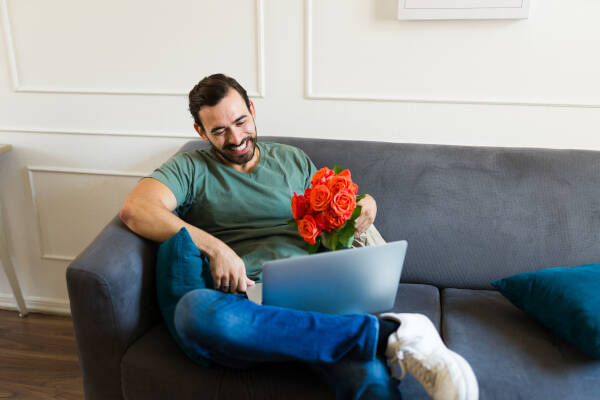 A man sitting on a couch with a laptop on his lap holds a bouquet of orange roses. He smiles joyfully while looking at the laptop screen.