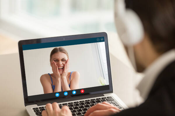 A man wearing headphones is using a laptop to video chat with a woman who is laughing and holding her face in joy. The laptop screen shows her delighted expression during the video call.