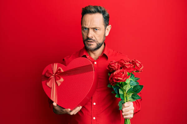 A man with a sarcastic expression holding a red heart-shaped gift box and roses against a red background.