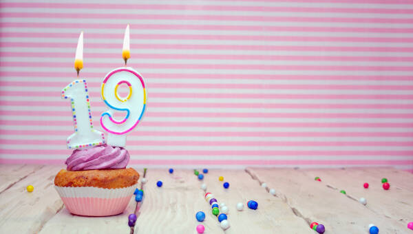 A muffin sits on a wooden surface with 19 candles atop it, against a white and pink striped background.