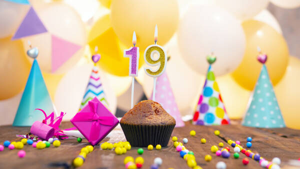 A muffin with '19' candles sits on a wooden surface with a gift box and party hats, with balloons in the background.