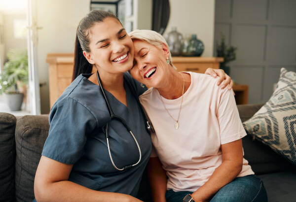 A senior woman hugs her doctor, sat in a living room, both smiling and laughing.