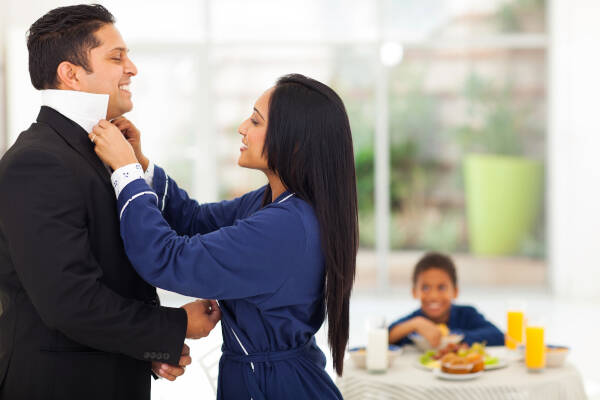 A wife helps her husband get dressed for work by fixing his tie, while their son watches from the breakfast table in the background.