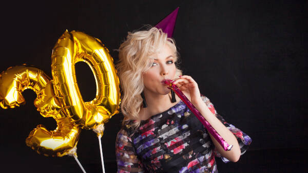 A woman blows a party horn as she holds golden '30' balloons against a black background.