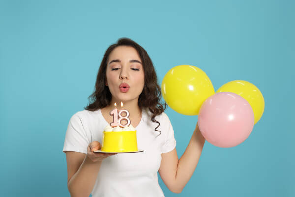 A woman blows out '18' candles on a cake while holding balloons in her other hand against a blue backdrop.