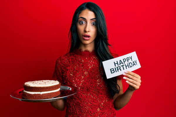 A woman holds up a piece of paper which says 'HAPPY BIRTHDAY' and holds a cake against a red backdrop.