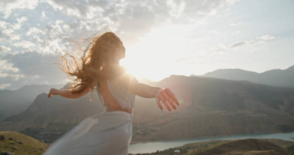 A woman in a white dress standing on top of a mountain at sunset, with raised hands as the wind blows.