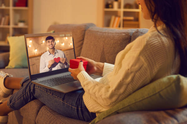 A woman sitting on a couch holds a small red gift box while video chatting with a man on her laptop screen. The man is holding a paper heart and smiling warmly.