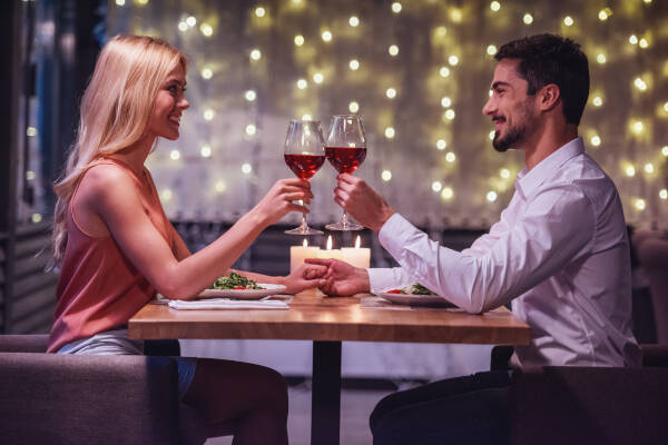 A young couple clinks glasses of wine, looking at each other and smiling during their date in a restaurant.