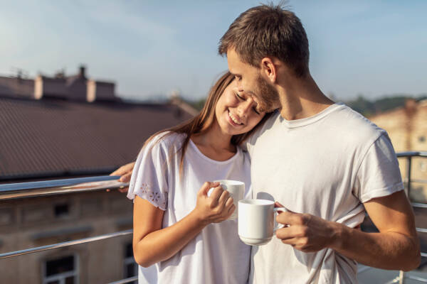 A young couple embraces on a rooftop in the morning, smiling as they hold cups of coffee.