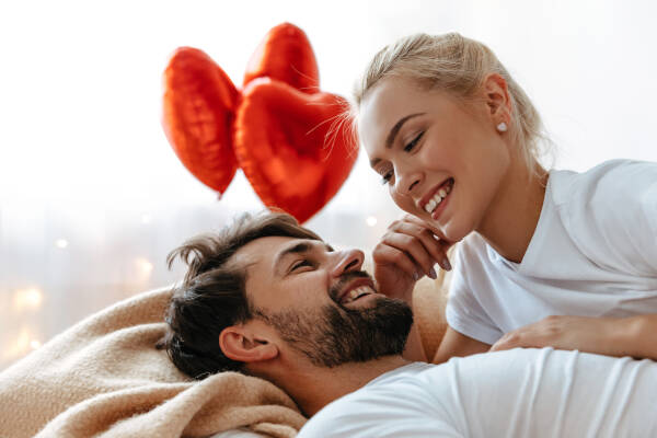 A young couple lies together on a bed, looking into each other's eyes and laughing, with three red heart shaped balloons in the background.