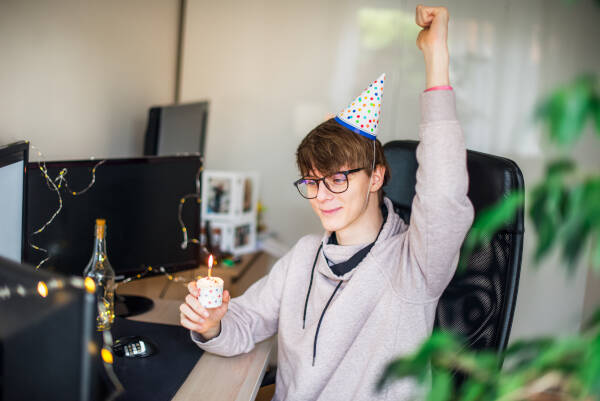 A young man sits at his desk with a party hat, cheering with a small cake adorned with one candle.