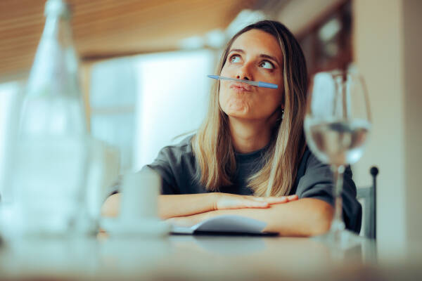 A young woman balances a pen under her nose while sitting at a table, looking up thoughtfully with a water bottle and glass nearby.