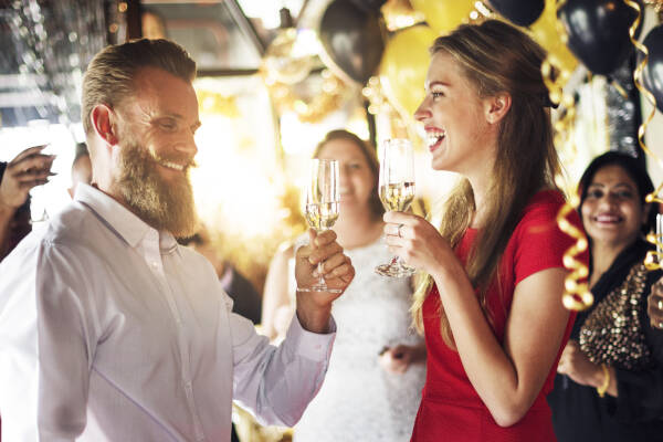 A young woman laughs while toasting her husband, with guests in the background and black and gold balloon decorations.