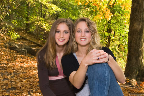 An 18-year-old big sister stands beside her 13-year-old little sister outdoors under trees in early fall.