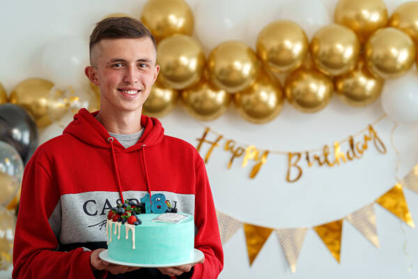 An 18-year-old boy stands with a cake at home, with golden banners and balloons in the background.