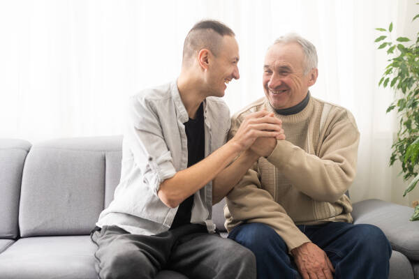 An elderly man sits on the sofa with his son, shaking hands and laughing together.