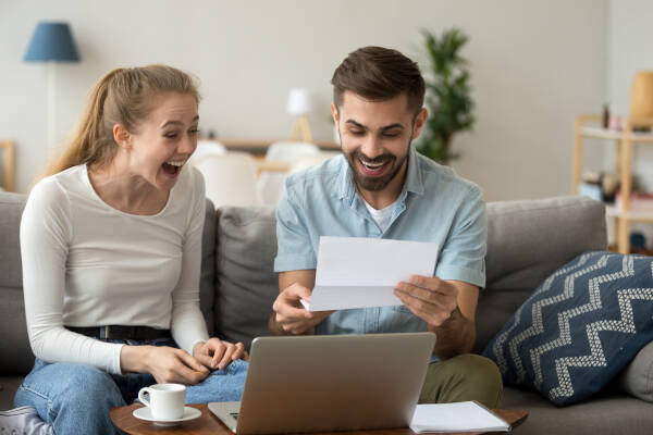 An excited and amazed couple, thrilled with joy, scream and rejoice as they read good news from a paper letter, sitting on the sofa at home.