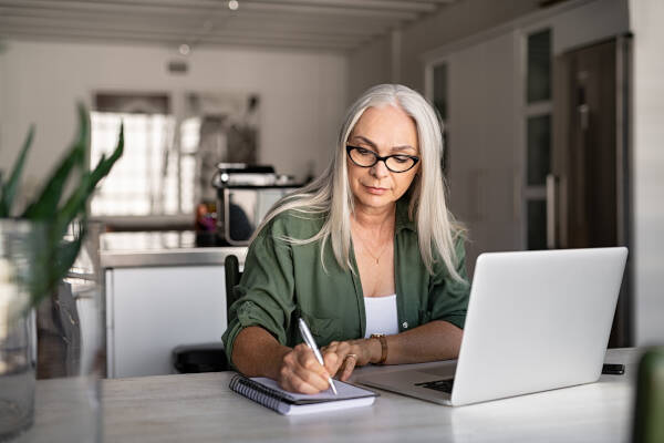 An older woman with white hair and glasses writes in a notebook at a table with a laptop, working in a well-lit kitchen.