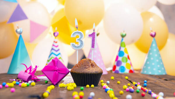 Cupcake with number '31' candles on a wooden table, next to a gift box and party hats, against a background of balloons.