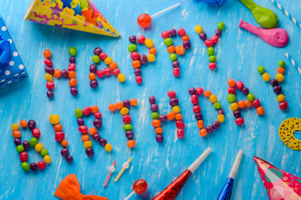 "HAPPY BIRTHDAY" spelled out in jelly beans on a blue background, surrounded by party hats, lollipops, and balloons.