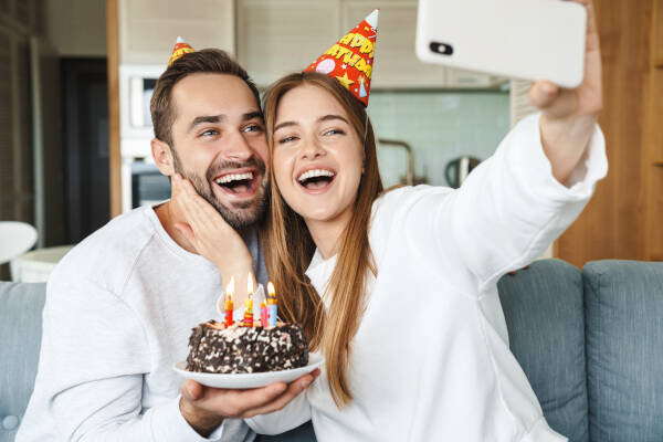 Happy, attractive young couple celebrating a birthday with a cake, sitting on a couch at home and taking a selfie.