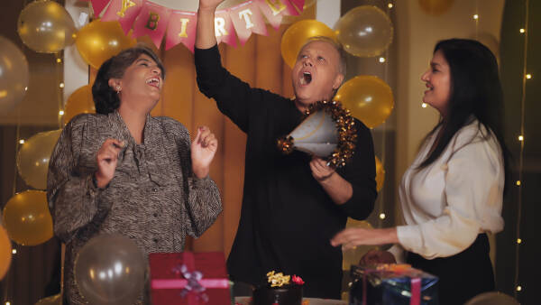 Indian elderly friends, smiling and joyful, dance to music at a night time indoor party, celebrating a birthday together in a festively decorated house.