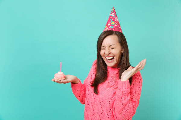 Laughing young woman in a knitted pink sweater and party hat holding a cupcake with a candle against a blue background.