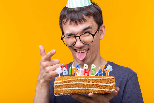 Man in spectacles pulls a funny expression while pointing at camera and holding a birthday cake, against orange background.