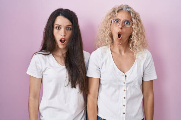 Mother and daughter stand against a pink backdrop with shocked expressions on their faces.