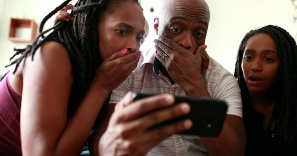 Parents and daughter staring at a smartphone with shocked expressions, holding their mouths.