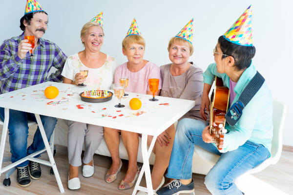 Seniors sit around a table wearing party hats, with one playing guitar and the others listening, all smiling.