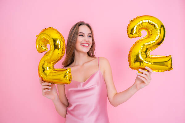 Smiling 22-year-old woman holds '22' balloons against a pink background.