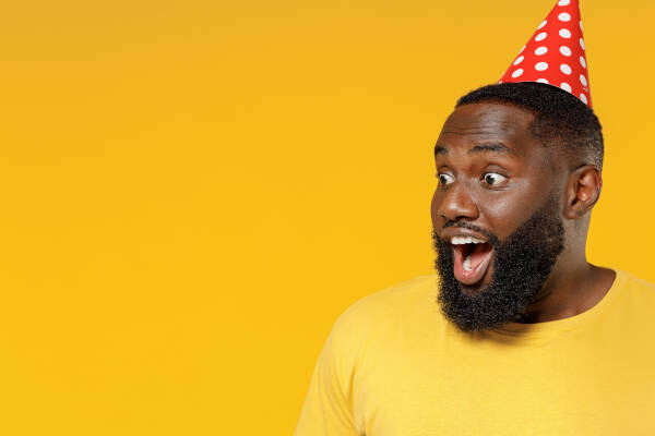 Surprised-looking man wearing a party hat stands in a yellow t-shirt against a yellow background.