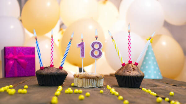 Three cupcakes, each with candles, on a wooden table with a pink gift box and balloons in the background.