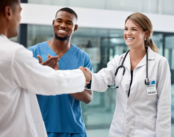 Three smiling doctors at a medical facility shake hands, expressing gratitude, congratulations, or a warm welcome.