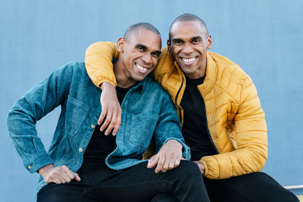 Twin brothers with arms around each other, smiling and posing for a photo against a blue background.