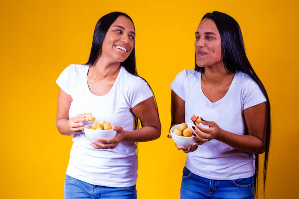 Twin sisters laughing together as they stand and eat food against a yellow background.