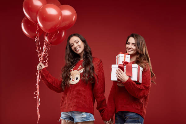 Twin sisters stand against a red backdrop; one holds gift boxes, and the other holds red balloons.