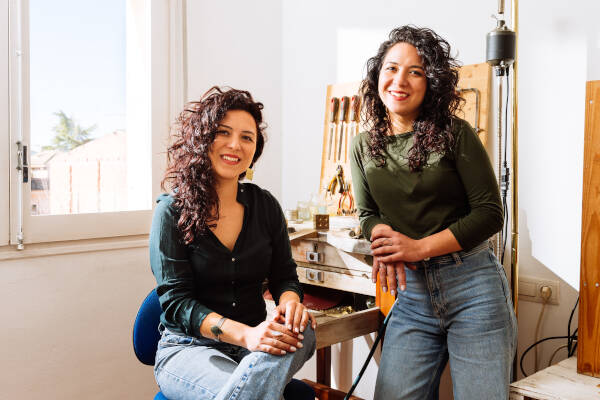 Two cheerful twin sisters indoors, posing for a photo with tools in the background.