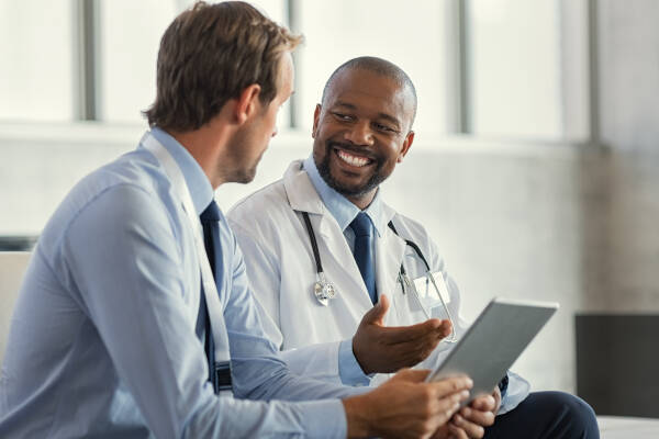 Two mature smiling male doctors sitting down and talking while one is holding a digital tablet, the other is smiling.